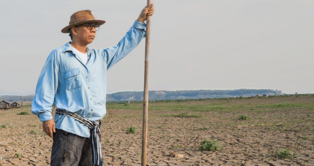man in a light blue shirt and brown hat stands holding a long staff against a clear sky backdrop